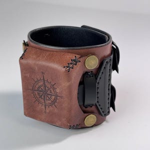 May include: A wide brown leather cuff bracelet featuring a square-shaped centre panel embossed with a black compass rose design. The cuff includes black leather accents, dark stitching, and two circular brass-toned metal studs on the front.