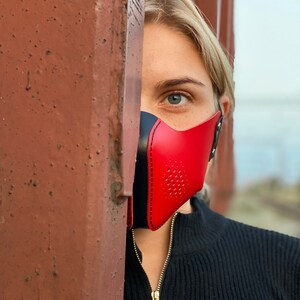 May include: A person wearing a red and black leather face mask with a perforated design. The mask is covering the person's mouth and nose. The person is looking at the camera.