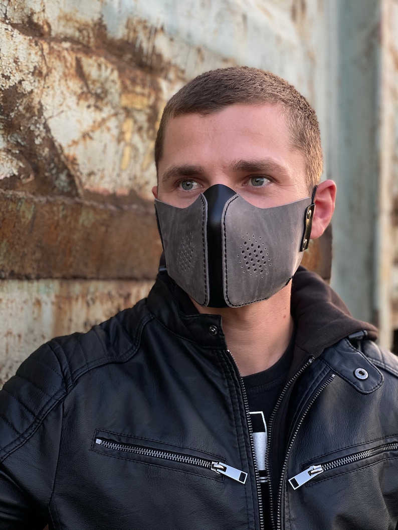 May include: A gray leather face mask with a black leather accent and a perforated design. The mask has a strap that goes around the ears.
