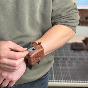 May include: Brown leather wrist-mounted phone case with a compass rose design. The case is secured with a snap closure and is designed to hold a smartphone. The person is wearing a green hoodie.