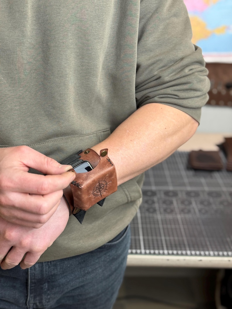 May include: Brown leather wrist-mounted phone case with a compass rose design. The case is secured with a snap closure and is designed to hold a smartphone. The person is wearing a green hoodie.