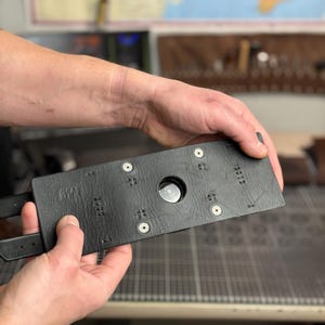 May include: A black leather belt buckle with a rectangular design. The buckle has a circular opening in the center and is secured with metal rivets. The belt has two straps.