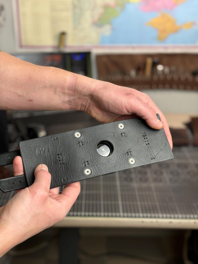May include: A black leather belt buckle with a rectangular design. The buckle has a circular opening in the center and is secured with metal rivets. The belt has two straps.