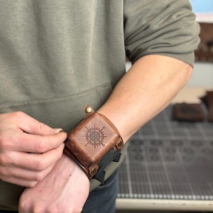May include: A brown leather wrist cuff with a compass rose design. The cuff is secured with snaps and a black strap. The image shows the cuff being fastened on a person's wrist. The background is blurred.