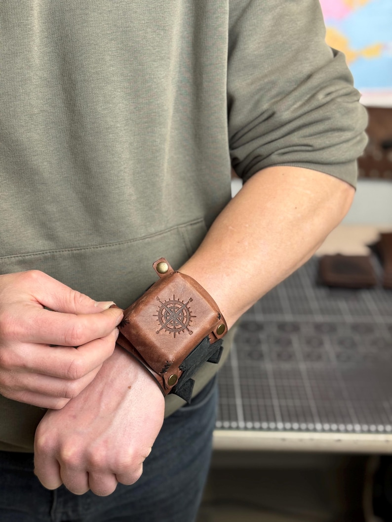 May include: A brown leather wrist cuff with a compass rose design. The cuff is secured with snaps and a black strap. The image shows the cuff being fastened on a person's wrist. The background is blurred.
