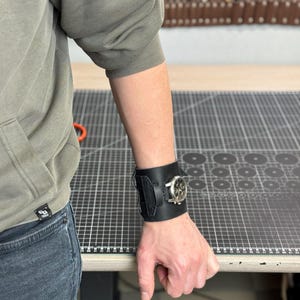 May include: A black leather watch cuff with a silver watch face. The cuff is wide, with a textured design and a buckle closure. The watch face has a silver bezel and a light-colored dial. The cuff is worn on a person's wrist.