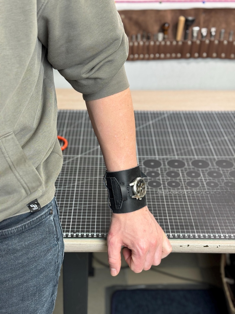 May include: A black leather watch cuff with a silver watch face. The cuff is wide, with a textured design and a buckle closure. The watch face has a silver bezel and a light-colored dial. The cuff is worn on a person's wrist.