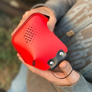 May include: Red leather case with a black strap and a small hole pattern. The case has black stitching around the edges.