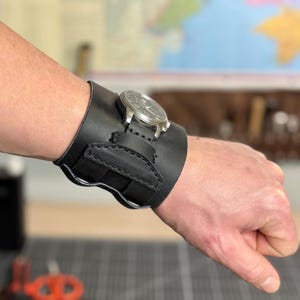 May include: A black leather watch cuff with a silver watch face. The cuff is wide, with a curved design and a small pocket. The watch is secured to the cuff with a strap. The watch cuff is worn on a wrist.