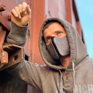 May include: A person wearing a gray hooded sweatshirt with a black leather face mask with a perforated design. The mask has a strap that goes around the back of the head. The person is standing in front of a rusty metal surface.