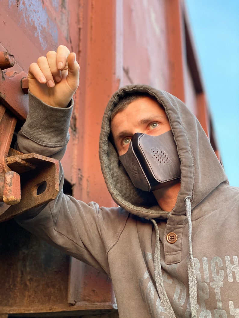 May include: A person wearing a gray hooded sweatshirt with a black leather face mask with a perforated design. The mask has a strap that goes around the back of the head. The person is standing in front of a rusty metal surface.