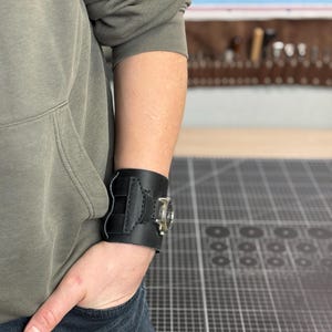 May include: Black leather wrist cuff with a silver-toned watch face. The cuff features decorative stitching and a buckle closure. The watch face has a clear glass cover. The cuff is worn on the wrist.