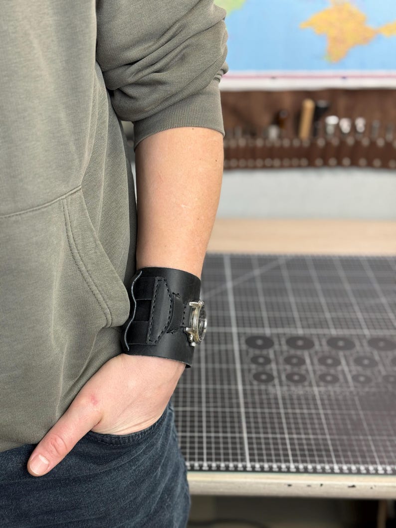 May include: Black leather wrist cuff with a silver-toned watch face. The cuff features decorative stitching and a buckle closure. The watch face has a clear glass cover. The cuff is worn on the wrist.