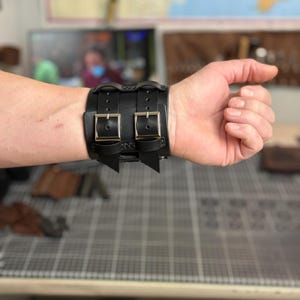 May include: Black leather wrist cuff with two silver-colored buckles. The cuff is secured with two straps and has decorative stitching. The cuff is worn on a wrist.