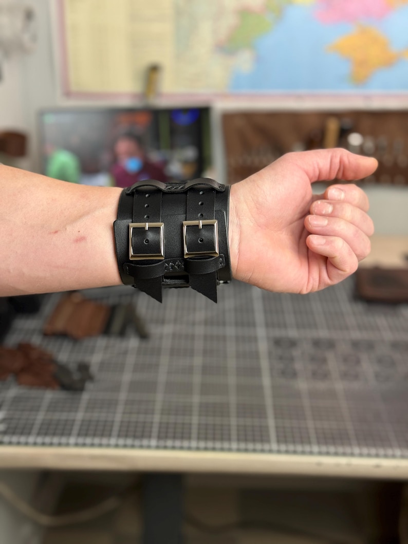 May include: Black leather wrist cuff with two silver-colored buckles. The cuff is secured with two straps and has decorative stitching. The cuff is worn on a wrist.