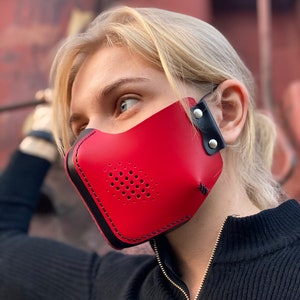 May include: A red leather face mask with black stitching and a black adjustable strap. The mask has a perforated design for breathability.