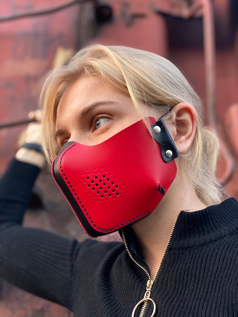 May include: A red leather face mask with black stitching and a black adjustable strap. The mask has a perforated design for breathability.