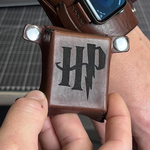 May include: A small, square, brown leather accessory with silver-tone metal snaps at the top corners. The front surface features a black engraved logo with the letters HP, where the letter P is stylized with a lightning bolt shape.