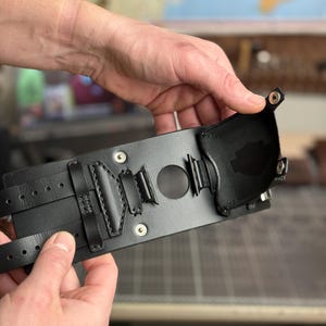 May include: A black leather wrist cuff with multiple straps and decorative stitching. The cuff features a central circular opening and is held by hands. The design includes metal rivets and a small, attached pouch.