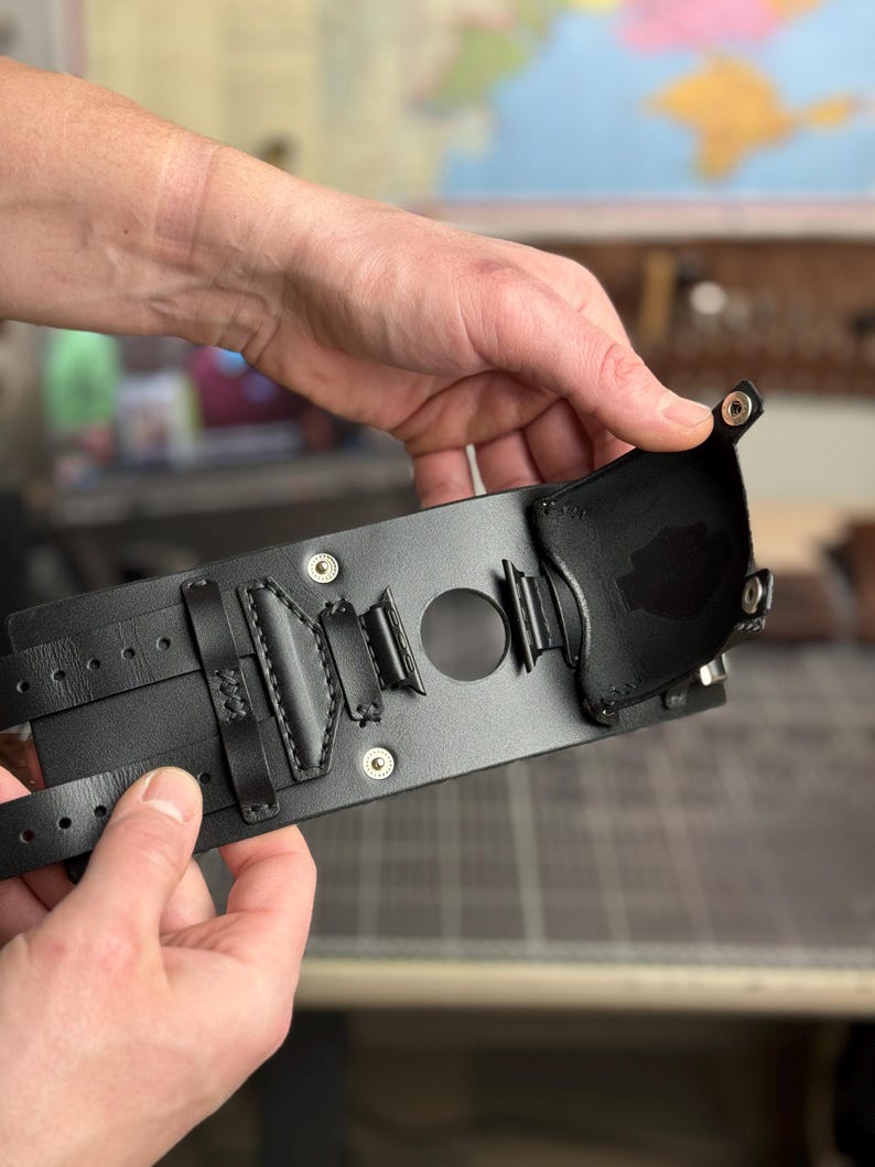 May include: A black leather wrist cuff with multiple straps and decorative stitching. The cuff features a central circular opening and is held by hands. The design includes metal rivets and a small, attached pouch.