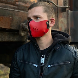 May include: A red leather face mask with black accents and a perforated design. The mask has adjustable straps and is worn by a person with a black leather jacket.