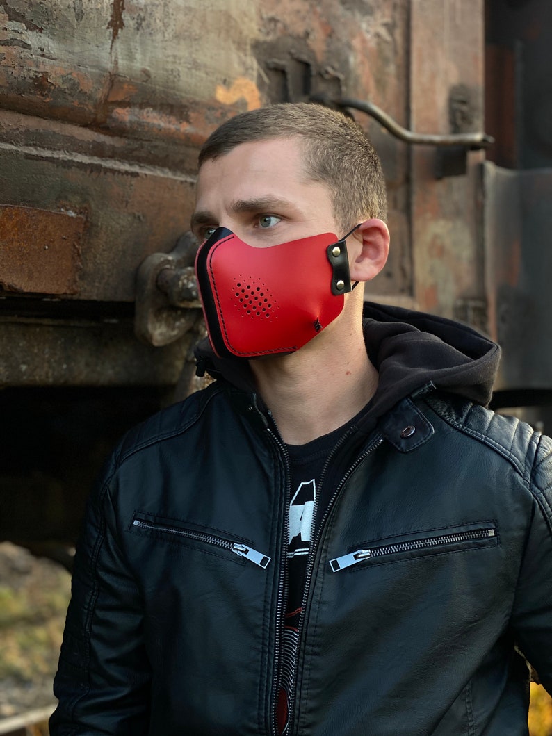 May include: A red leather face mask with black accents and a perforated design. The mask has adjustable straps and is worn by a person with a black leather jacket.