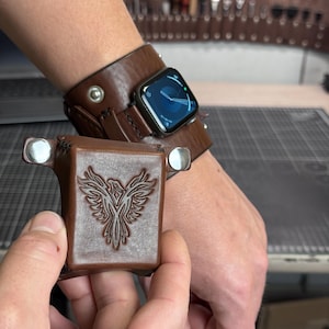 May include: A brown leather watch cuff with a dark smartwatch face and a matching brown leather square pouch featuring an embossed phoenix design, secured with silver-toned metal snaps on the sides and stitched edges.