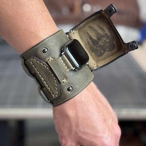 May include: An olive green leather cuff watch band featuring a wide strap with metal snap buttons and a hinged cover. The interior of the cover displays a stamped bear paw print design. The band secures a black smartwatch face in the center.