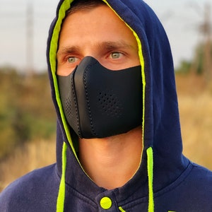 May include: A person wearing a black face mask with a navy blue hoodie with a lime green lining. The hoodie has a lime green drawstring and buttons.
