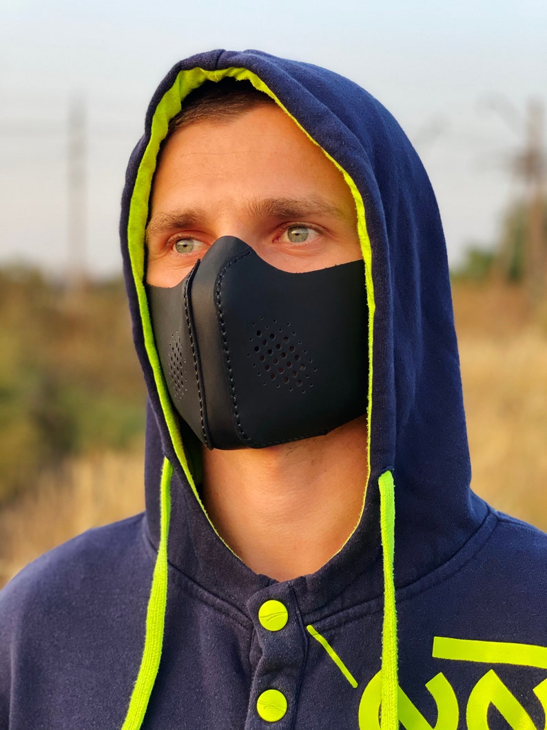 May include: A person wearing a black face mask with a navy blue hoodie with a lime green lining. The hoodie has a lime green drawstring and buttons.
