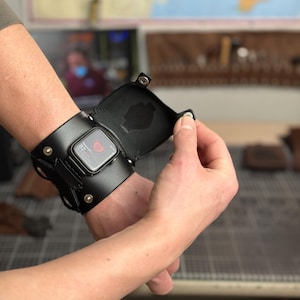 May include: Black leather Apple Watch cuff with a hinged cover, secured by metal rivets. The watch face displays a digital interface. The cuff wraps around the wrist, showcasing a blend of functionality and style.