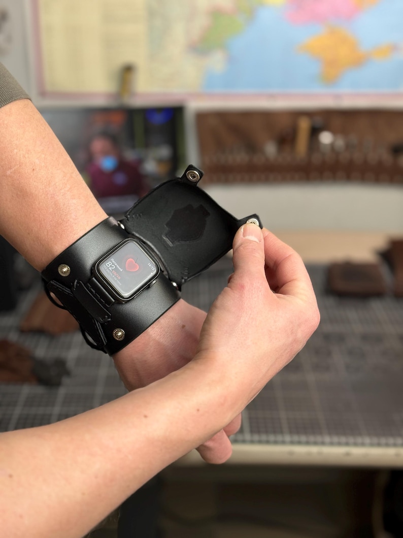 May include: Black leather Apple Watch cuff with a hinged cover, secured by metal rivets. The watch face displays a digital interface. The cuff wraps around the wrist, showcasing a blend of functionality and style.