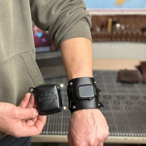 May include: Black leather Apple Watch cuff with silver rivets and a matching square case with the Harley-Davidson logo. The watch cuff is worn on the wrist, and the case is held in the hand. The watch face is visible.