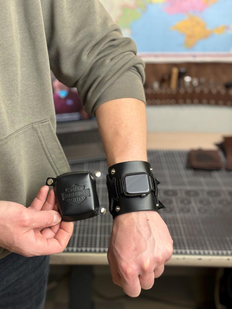 May include: Black leather Apple Watch cuff with silver rivets and a matching square case with the Harley-Davidson logo. The watch cuff is worn on the wrist, and the case is held in the hand. The watch face is visible.