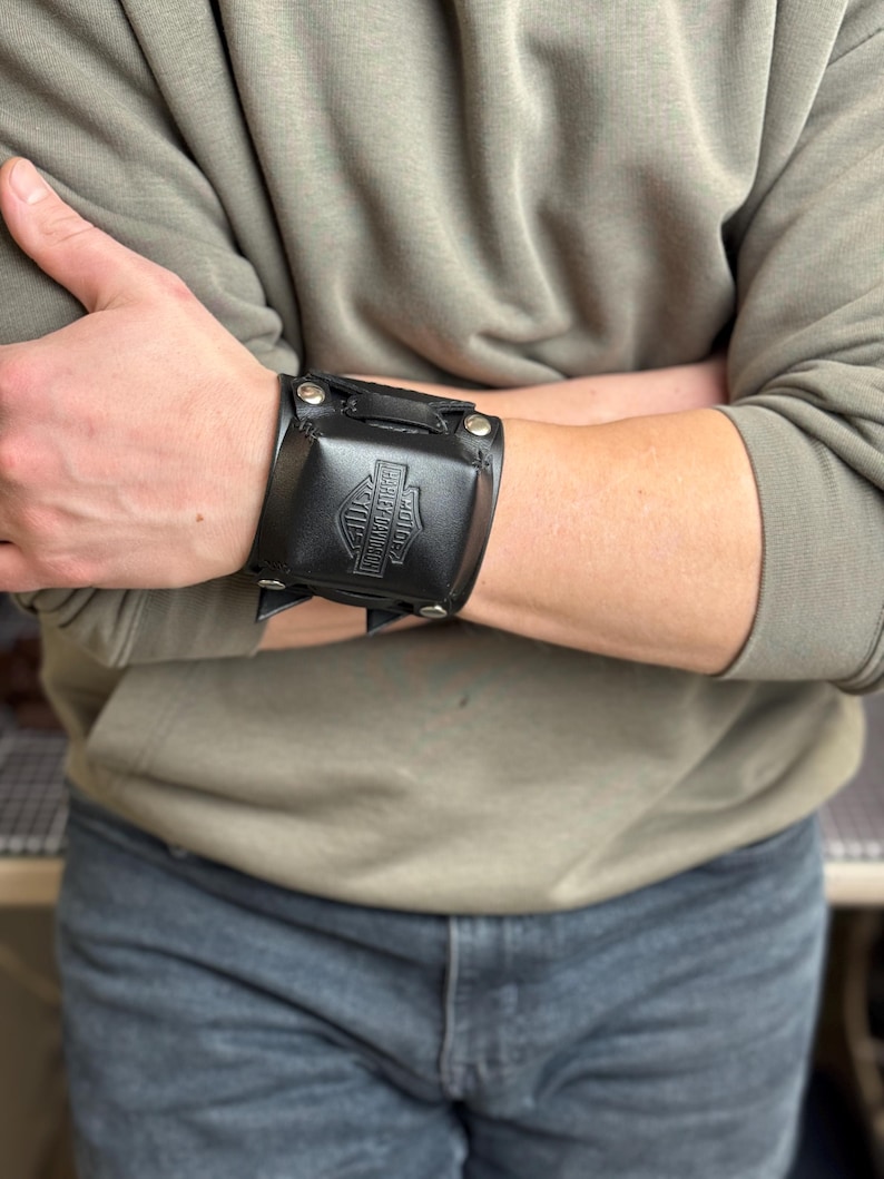 May include: Black leather wrist cuff featuring the Harley-Davidson logo. The cuff is secured with metal snaps and has a rectangular design. The cuff is worn on the wrist. The background is a person wearing a green sweatshirt and blue jeans.
