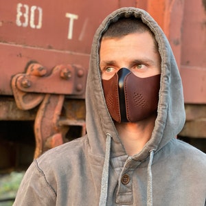 May include: A person wearing a gray hooded sweatshirt with the text "BETTER RICH" printed on the front. They are wearing a brown leather face mask with black accents and a perforated design.