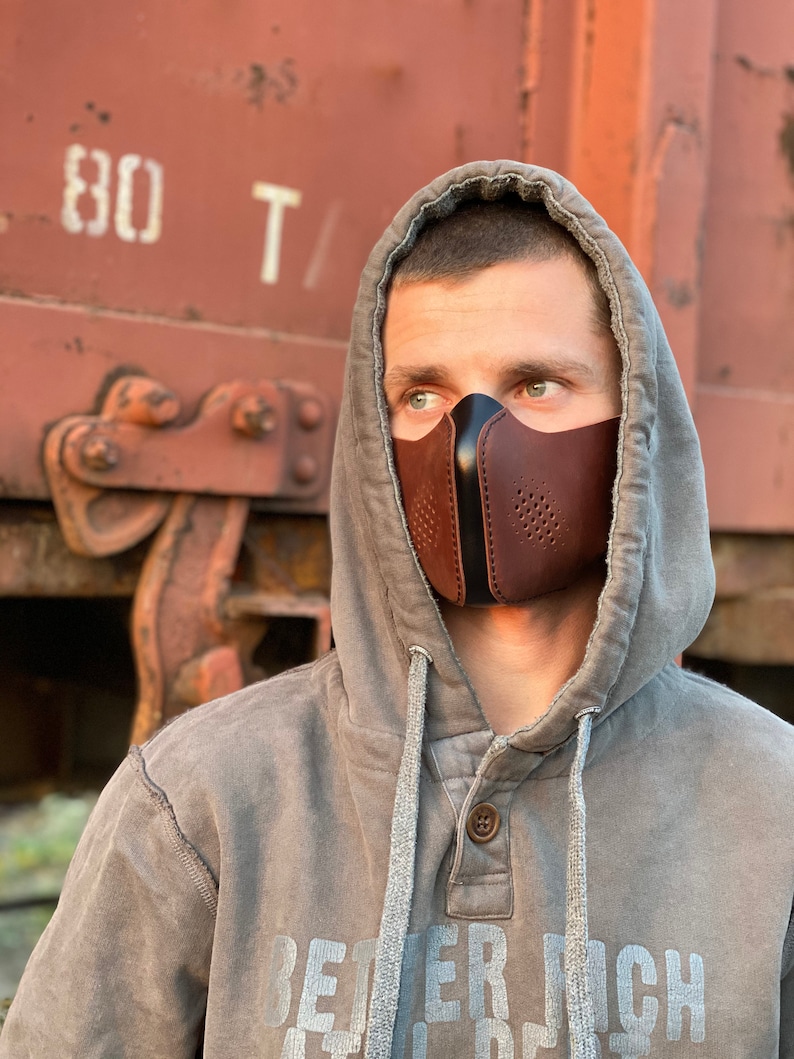 May include: A person wearing a gray hooded sweatshirt with the text "BETTER RICH" printed on the front. They are wearing a brown leather face mask with black accents and a perforated design.