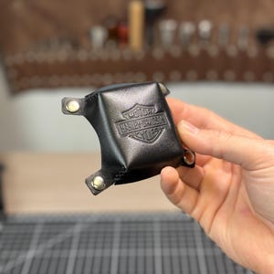 May include: A small black leather valet tray with a square shape and folded corners secured by silver-coloured metal snaps. The centre of the tray features an embossed Harley-Davidson Motor Cycles shield logo. The leather has a smooth finish.