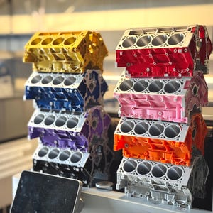 May include: Stacks of colorful engine blocks in yellow, blue, purple, black, red, pink, orange, and silver. Each block has multiple circular openings. The blocks are stacked on a shelf in a workshop setting.