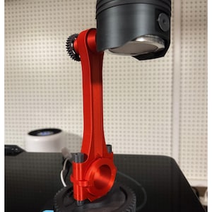 May include: A unique desk lamp featuring a black piston head and a red connecting rod. The lamp is mounted on a black gear base. The design incorporates automotive components, creating an industrial aesthetic.