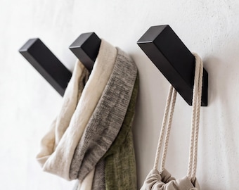 May include: Three matte black wall-mounted hooks with a square profile. One hook holds a folded beige and olive green scarf, while another hook supports a small beige drawstring bag with braided rope handles. The hooks are angled upward.