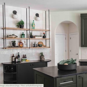 May include: A kitchen featuring a black metal ceiling-mounted shelving unit with three light wood shelves. The shelves hold various decorative items, including glass bottles, small potted plants, a fish figurine, and books against a white tiled wall.