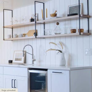 May include: Two-tier open shelving unit with black metal frames and light wood shelves mounted on a white vertical paneled wall. The shelves hold glassware, wooden bowls, a cutting board, and decorative items above a white kitchen counter.
