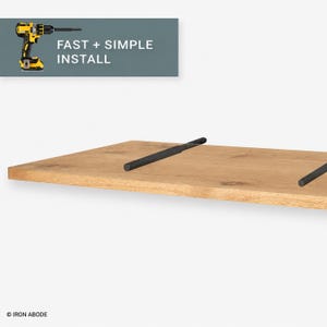 May include: A light brown wooden floating shelf with two black metal mounting rods extending from the surface. A graphic in the upper left corner shows a yellow and black power drill with the text FAST + SIMPLE INSTALL.
