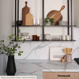 May include: A black metal shelving unit mounted on a wall, featuring two tiers holding wooden cutting boards, ceramic bowls, glassware, and small potted plants. Below, a marble countertop holds a black vase with greenery and a utensil holder.