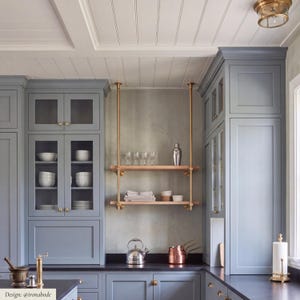 Può includere: Una cucina con armadietti blu-grigi con ante a vetro, una mensola in legno a due livelli sospesa da aste in ottone, un bollitore in acciaio inossidabile, pentole in rame e un portarotolo su un piano di lavoro scuro sotto un soffitto bianco.