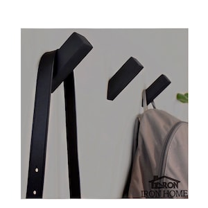 May include: Three black wooden wall hooks mounted on a white wall. A black leather strap hangs on the leftmost hook, and a gray backpack hangs on the rightmost hook. The hooks are angled slightly upward. The text "IRON HOME" is visible in the bottom right corner of the image.
