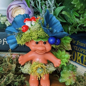 May include: A small troll doll with blue eyes and a big smile, wearing a crown of moss, blue flowers, and red mushrooms. The doll is wearing a green moss skirt with a yellow flower.