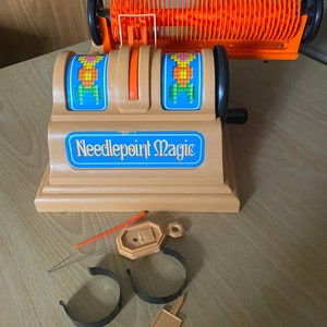 May include: A vintage needlepoint magic loom with orange and black plastic parts. The loom has two spinning wheels with colorful needlepoint designs. The loom is on a wooden surface with a needle, two black plastic hoops, and a small orange tag.