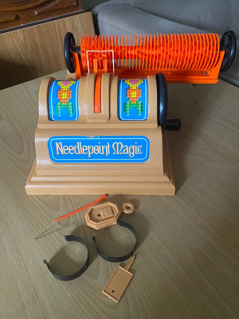 May include: A vintage needlepoint magic loom with orange and black plastic parts. The loom has two spinning wheels with colorful needlepoint designs. The loom is on a wooden surface with a needle, two black plastic hoops, and a small orange tag.